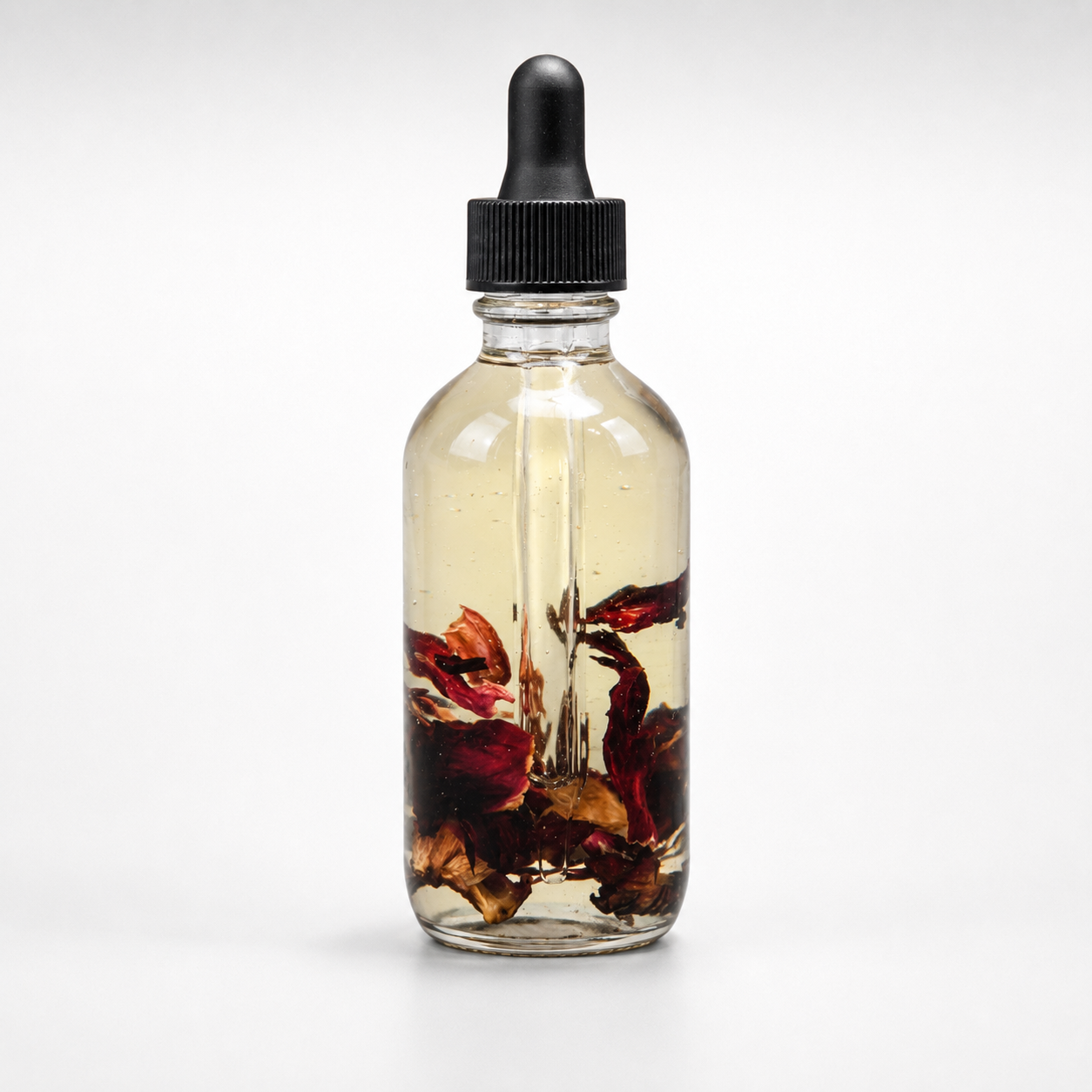 A clear glass dropper bottle with a black cap, filled with a light amber liquid and containing visible dried red and brown botanical pieces, photographed on a clean white background.